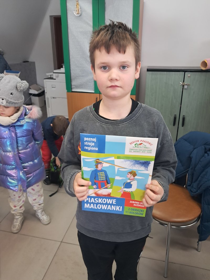 Zdjęcie przedstawia dziecko trzymające w rękach „piaskowe malowidło” z którego można namalować strój ludowy na kartce papieru.  