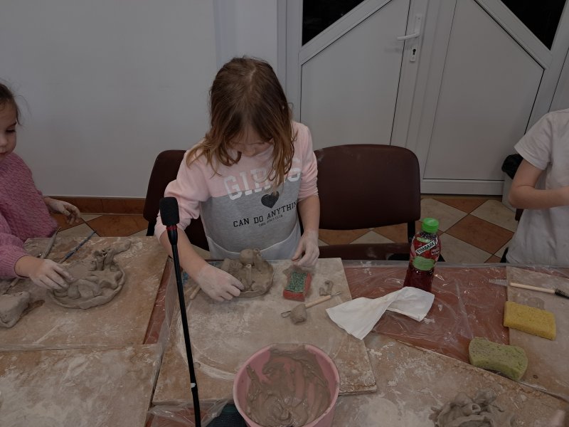 Zdjęcie przedstawia uczestniczkę zajęć z ceramiki siedzącą na krześle przy stoliku w trakcie lepienia z gliny  