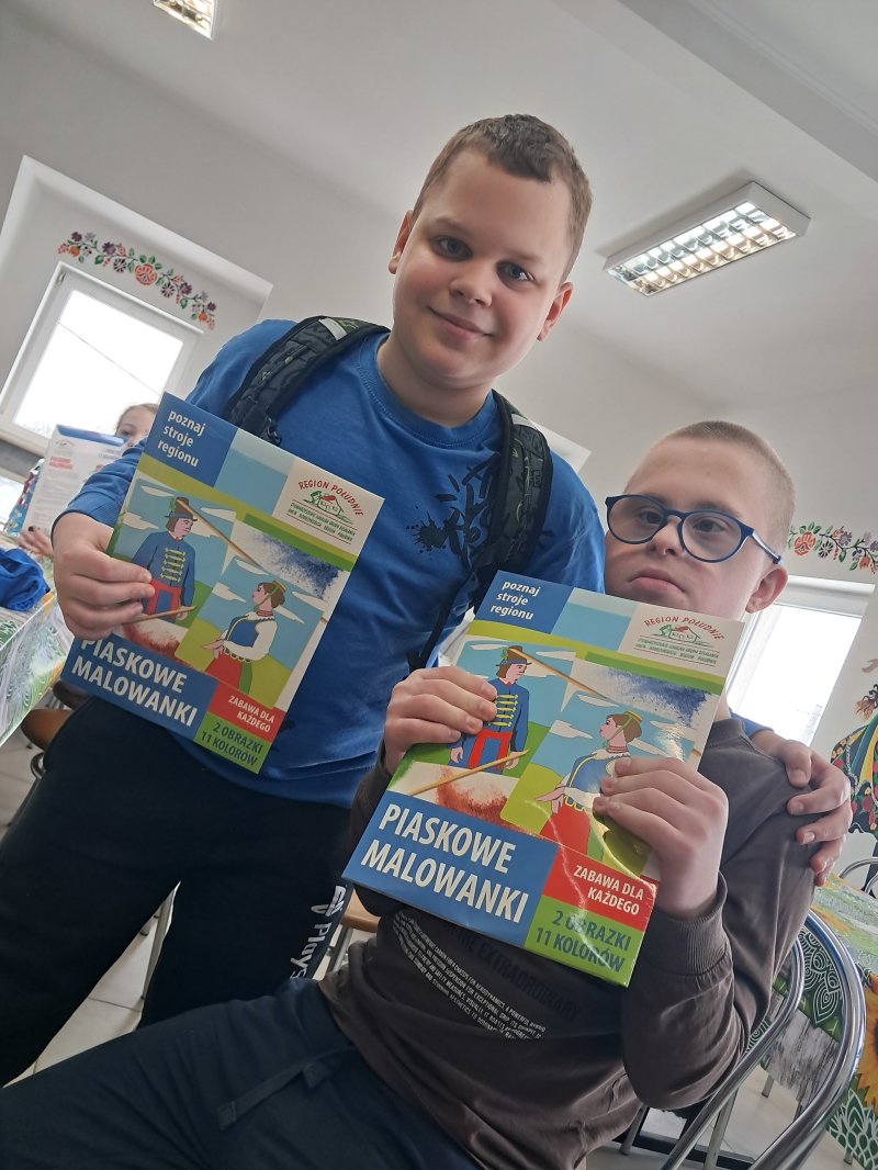 Zdjęcie przedstawia dzieci trzymające w rękach „piaskowe malowidło” z którego można namalować strój ludowy na kartce papieru.  