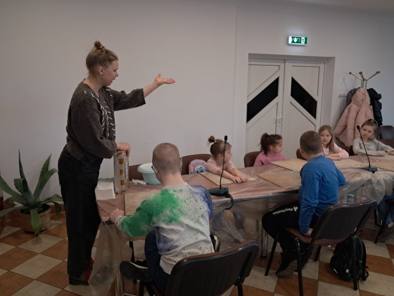 Zdjęcie przedstawia uczestników zajęć siedzących na krzesłach przy stoliku w trakcie warsztatów z ceramiki w towarzystwie instruktorki prowadzącej warsztaty, która gestykuluje rękami coś tłumacząc dzieciom . Przed dziećmi na stolikach leżą przybory do pracy.   