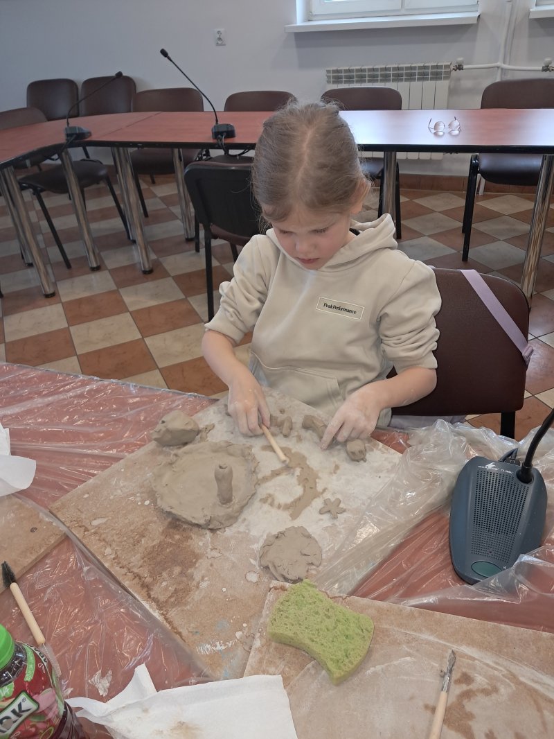 Zdjęcie przedstawia uczestniczkę zajęć z ceramiki siedzącą na krześle przy stoliku w trakcie lepienia z gliny  