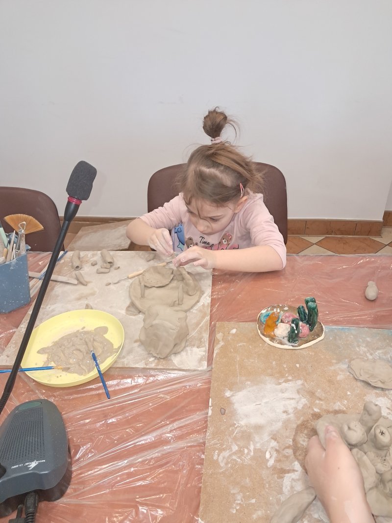 Zdjęcie przedstawia uczestniczkę zajęć z ceramiki siedzącą na krześle przy stoliku w trakcie lepienia z gliny  