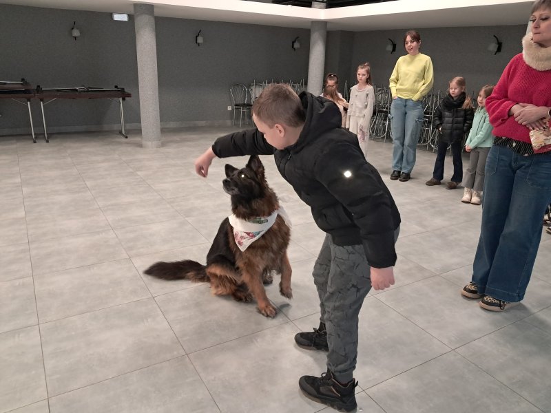 Zdjęcie przedstawia psa w towarzystwie swojego opiekuna i uczestnika zajęć podającego psu pokarm w sali, w której odbywały się zajęcia. W dali widać innych uczestników spotkania  