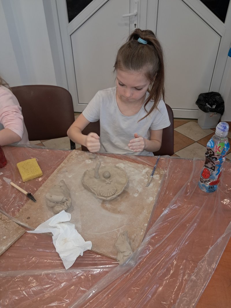 Zdjęcie przedstawia uczestniczkę zajęć z ceramiki siedzącą na krześle przy stoliku w trakcie lepienia z gliny  