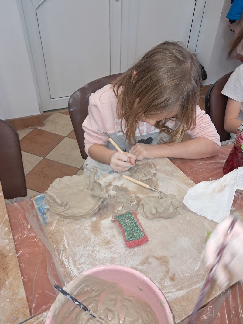 Zdjęcie przedstawia uczestniczkę zajęć z ceramiki siedzącą na krześle przy stoliku w trakcie lepienia z gliny  