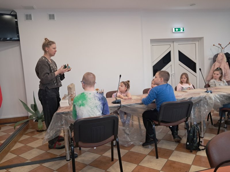 Zdjęcie przedstawia uczestników zajęć siedzących na krzesłach przy stoliku w trakcie warsztatów z ceramiki w towarzystwie instruktorki prowadzącej warsztaty, która trzyma w rękach figurkę ulepioną z gliny. Przed dziećmi na stolikach leżą przybory do pracy.   