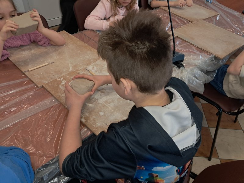 Zdjęcie przedstawia uczestnika zajęć z ceramiki siedzącego na krześle przy stoliku w trakcie zajęć i trzymającego w dłoni kostkę z gliny  