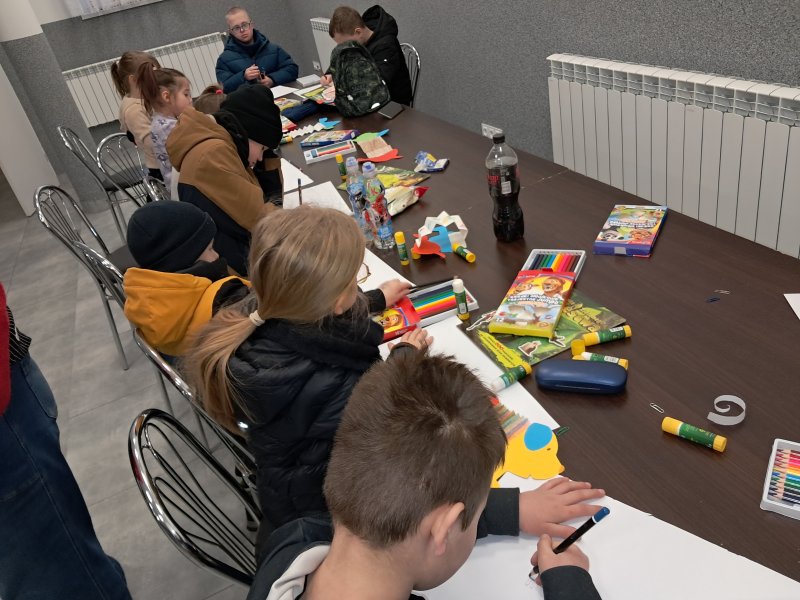Zdjęcie przedstawia uczestników zajęć siedzących na krzesłach przy stoliku w trakcie zajęć plastycznych  