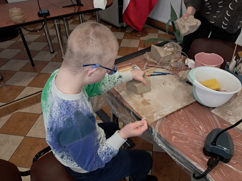 Zdjęcie przedstawia uczestnika zajęć z ceramiki siedzącego na krześle przy stoliku w trakcie zajęć i trzymającego w dłoni kostkę z gliny.  