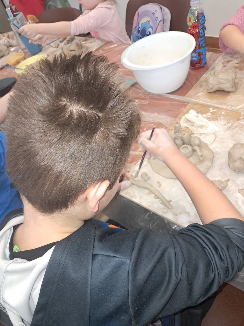 Zdjęcie przedstawia uczestnika zajęć z ceramiki siedzącą na krześle przy stoliku w trakcie lepienia z gliny  