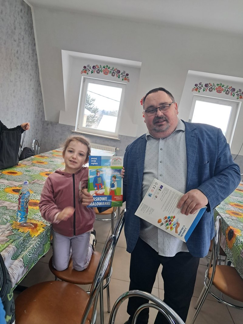 Zdjęcie przedstawia dziecko trzymające w rękach „piaskowe malowidło” z którego można namalować strój ludowy na kartce papieru. Razem z dziewczynką do zdjęcia pozuje dyrektor biblioteki.  