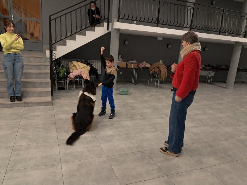 Zdjęcie przedstawia psa w towarzystwie swojego opiekuna i uczestnika zajęć w sali, w której odbywały się zajęcia.  