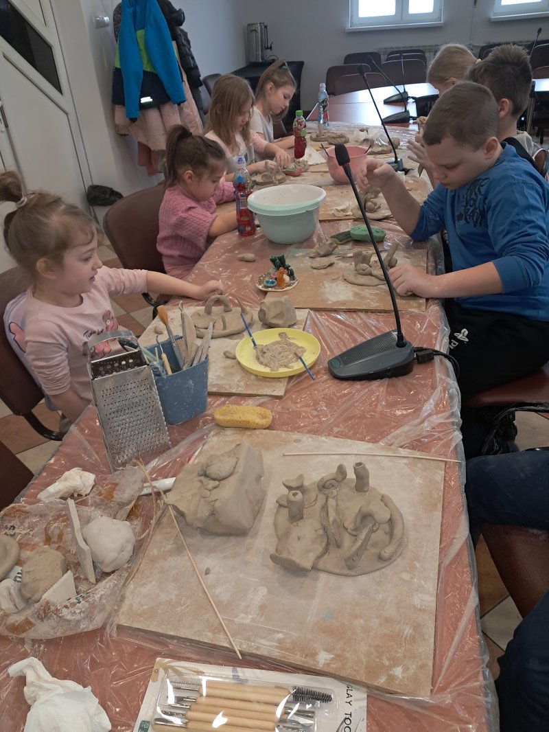 Zdjęcie przedstawia uczestników zajęć siedzących na krzesłach przy stoliku w trakcie warsztatów z ceramiki.  