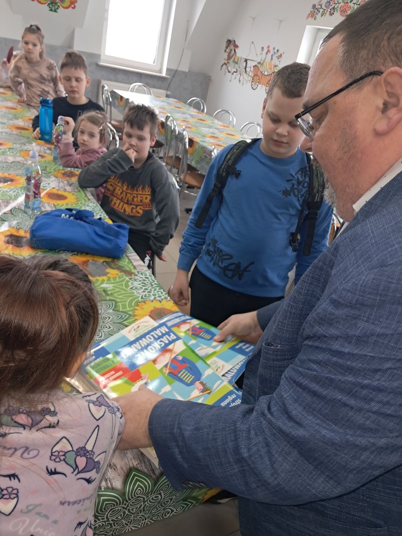 Na zdjęciu widać dyrektora biblioteki stojącego przy stoliku, trzymającego w ręku „piaskowe malowidło”, potrzebne do pracy plastycznej. Dookoła stoją dzieci.  