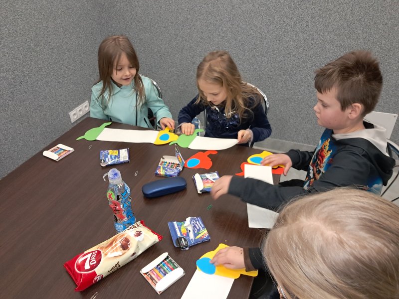 Zdjęcie przedstawia uczestników zajęć siedzących na krzesłach przy stoliku w trakcie zajęć plastycznych  