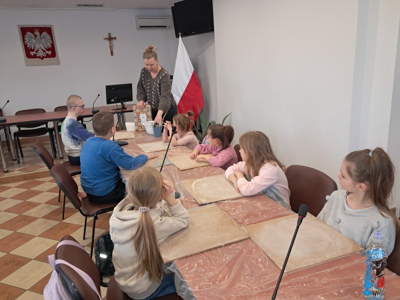 Zdjęcie przedstawia uczestników zajęć siedzących na krzesłach przy stoliku w trakcie warsztatów z ceramiki w towarzystwie instruktorki prowadzącej warsztaty. Przed dziećmi na stolikach leżą przybory do pracy  