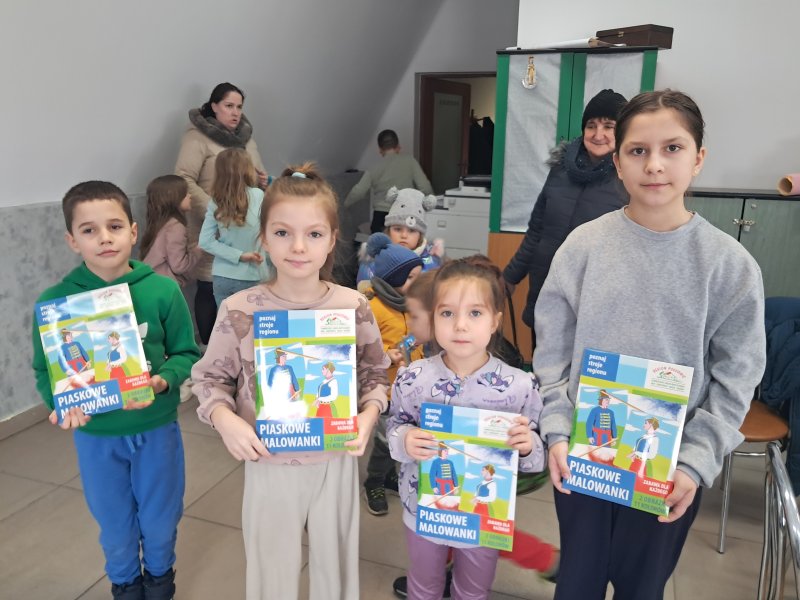 Zdjęcie przedstawia dzieci trzymające w rękach „piaskowe malowidło” z którego można namalować strój ludowy na kartce papieru.  
