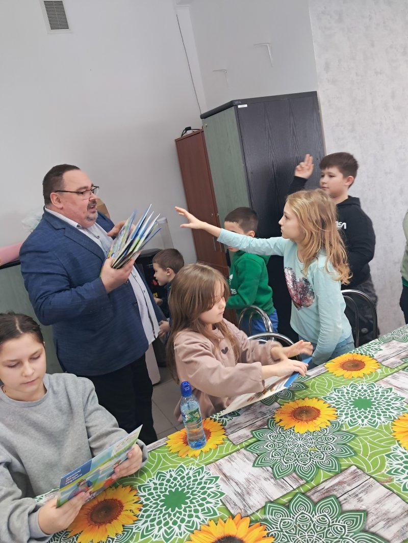 Na zdjęciu widać dyrektora biblioteki stojącego przy stoliku, wręczającego dzieciom „piaskowe malowidło”, potrzebne do pracy plastycznej.   