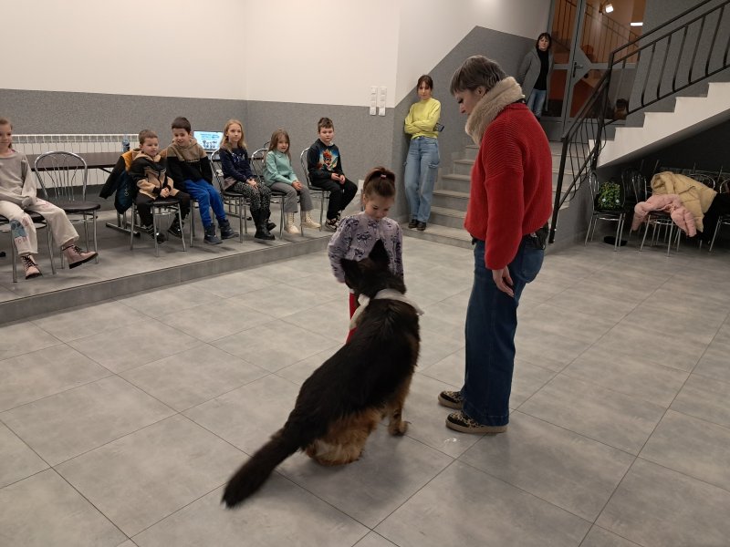 Zdjęcie przedstawia psa w towarzystwie opiekuna i uczestniczki zajęć w sali, w której odbywały się zajęcia. Na dalszym planie widać dzieci siedzące na krzesłach  