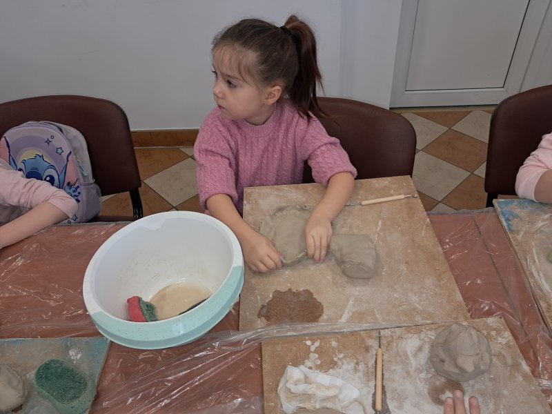 Zdjęcie przedstawia uczestniczkę zajęć z ceramiki siedzącą na krześle przy stoliku w trakcie lepienia z gliny  