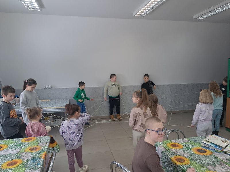 Na zdjęciu widać grupkę dzieci stojących w kręgu w Sali, w której odbywały się zajęcia. Dzieci trzymają w rękach nitki w trakcie zabawy w pajęczynę.  