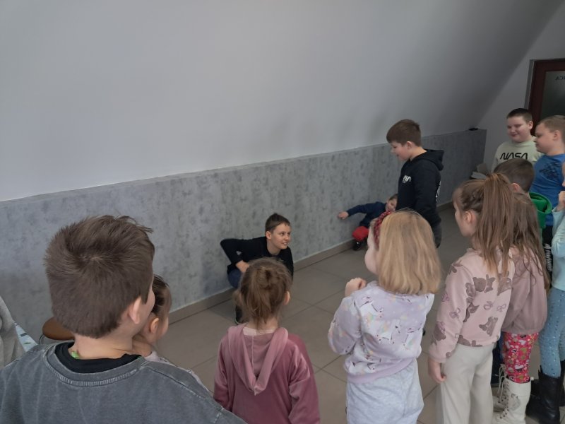 Na zdjęciu widać  wspierającego się na rękach na podłodze chłopca w trakcie gry w kalambury. Dookoła w sali, w której odbywały się zajęcia stoją dzieci.  