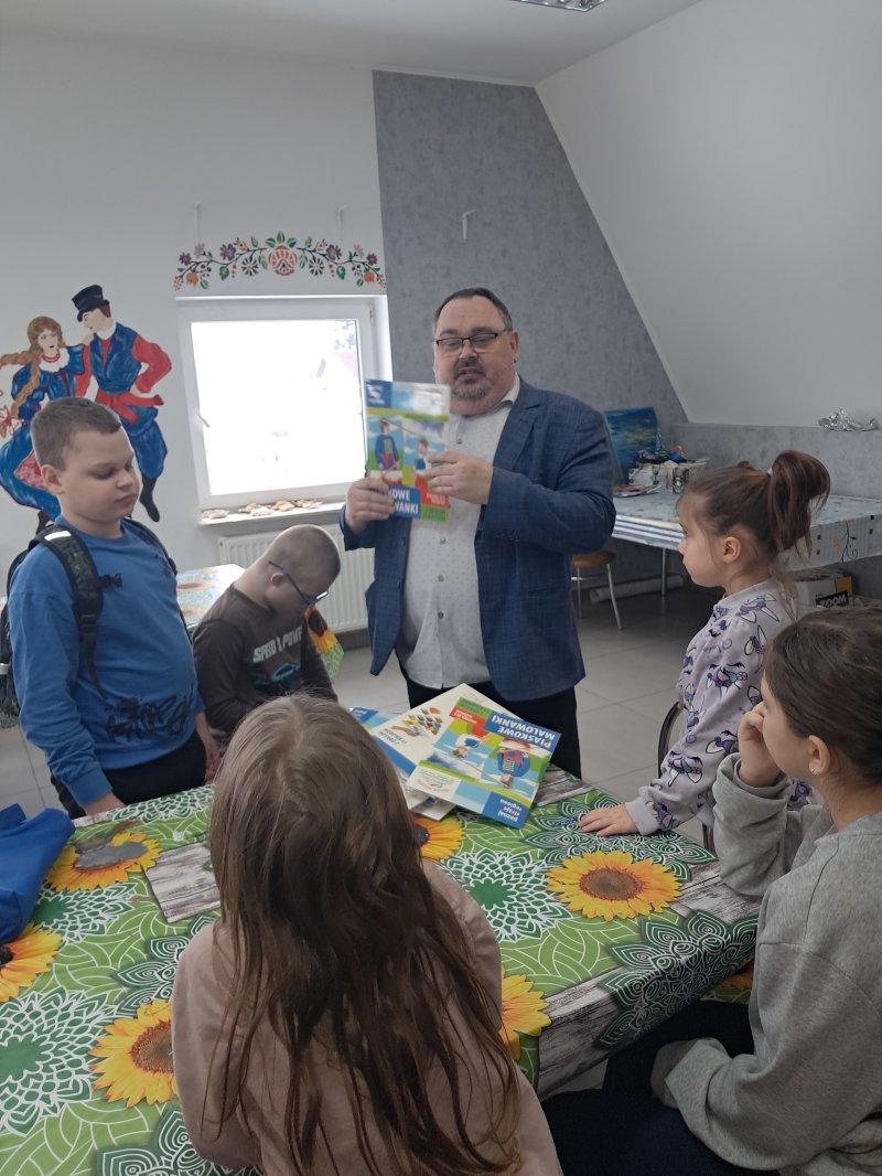 Na zdjęciu widać dyrektora biblioteki stojącego przy stoliku, trzymającego w ręku „piaskowe malowidło”, potrzebne do pracy plastycznej. Dookoła stoją dzieci.  