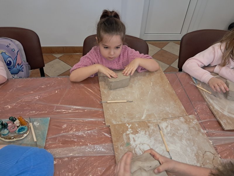 Zdjęcie przedstawia uczestniczkę zajęć z ceramiki siedzącą na krześle przy stoliku w trakcie zajęć i trzymającą w dłoni kostkę z gliny  