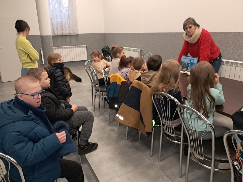 Zdjęcie przedstawia uczestników zajęć siedzących na krzesłach przy stoliku w sali, w której odbywają się zajęcia. Przed dziećmi stoi pani, która pokazuje im prezentację na komputerze  