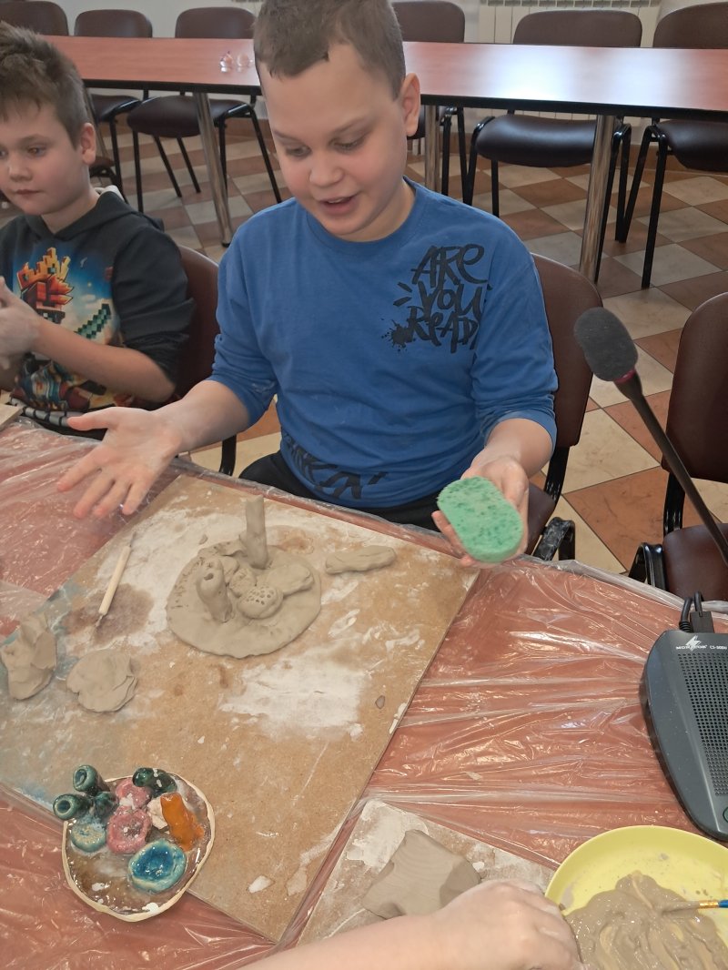 Zdjęcie przedstawia uczestnika zajęć z ceramiki siedzącą na krześle przy stoliku w trakcie lepienia z gliny  