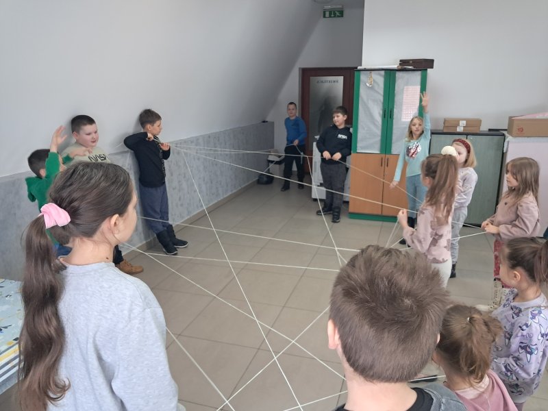 Na zdjęciu widać grupkę dzieci stojących w kręgu w Sali, w której odbywały się zajęcia. Dzieci trzymają w rękach nitki w trakcie zabawy w pajęczynę.  