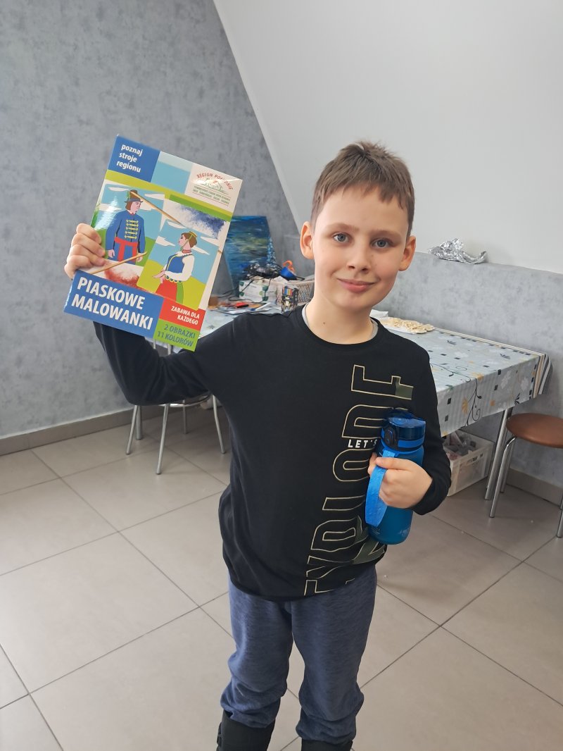 Zdjęcie przedstawia dziecko trzymające w rękach „piaskowe malowidło” z którego można namalować strój ludowy na kartce papieru.  