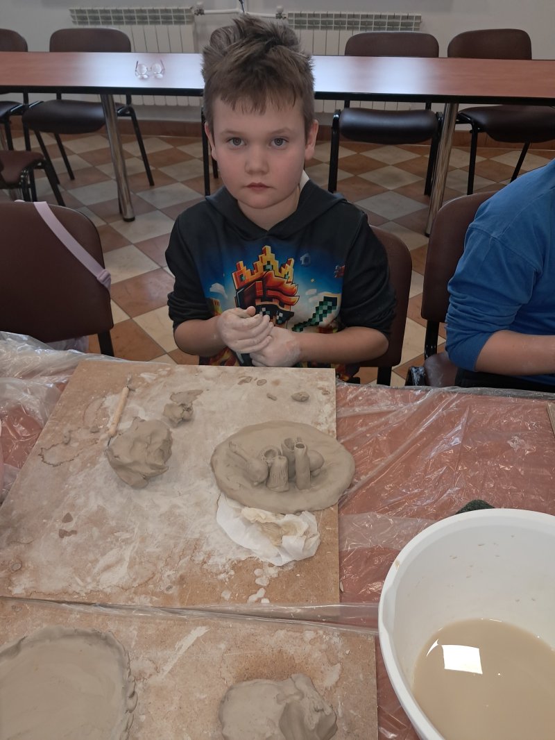 Zdjęcie przedstawia uczestnika zajęć z ceramiki siedzącą na krześle przy stoliku w trakcie lepienia z gliny  