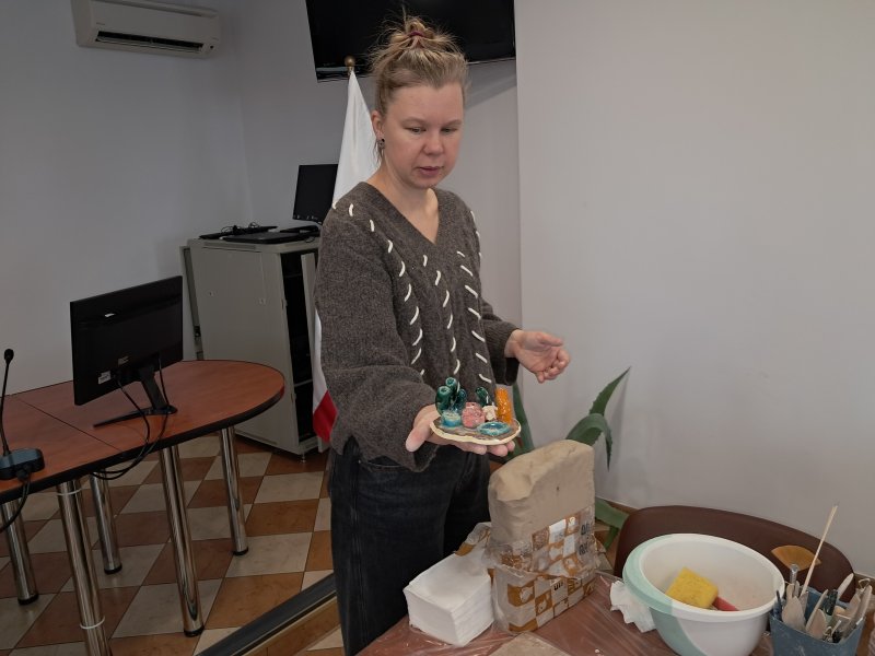 Zdjęcie przedstawia osobę prowadzącą warsztaty z ceramiki która trzyma w rękach gotową figurkę ulepioną i wypaloną z gliny  