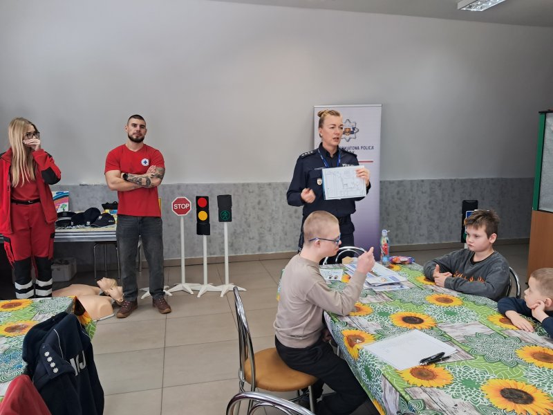 Zdjęcie przedstawia rzecznika prasowego policji Małgorzatę Małkińską stojącą w sali, w której odbywały się zajęcia i mówiącą do dzieci, które siedzą przy stoliku. W dali widać ratowników Lipnowskiego WOPR,  