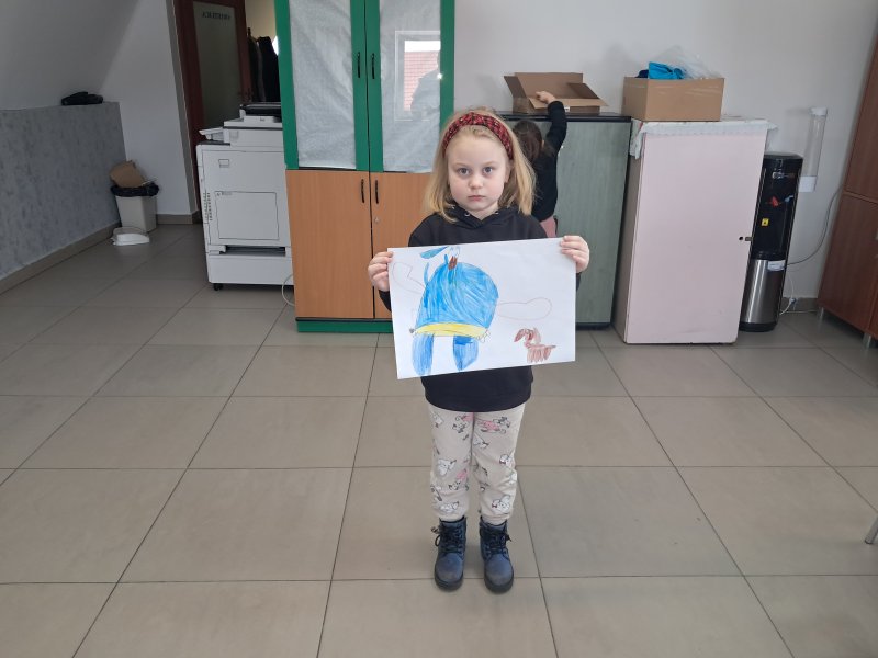 Zdjęcie przedstawia dziewczynkę stojącą i pozującą do zdjęcia z własnym rysunkiem  