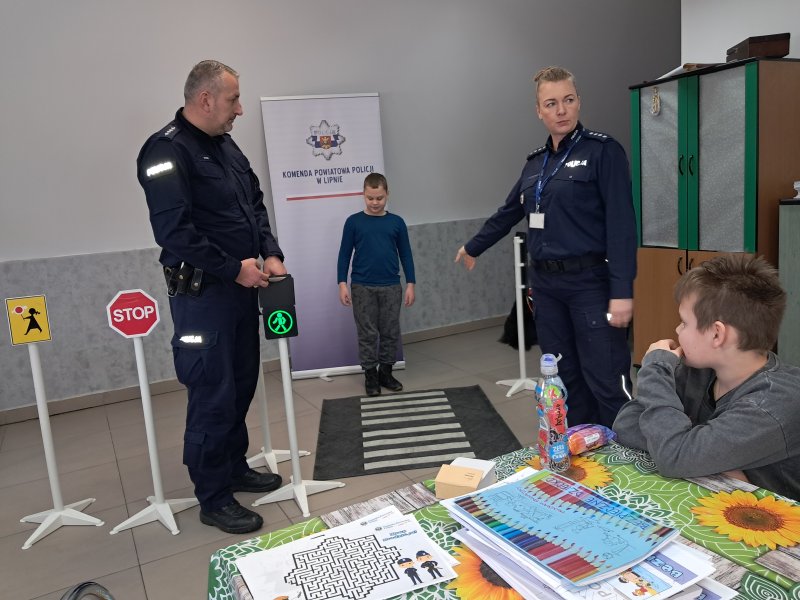 Zdjęcie przedstawia policjantów i uczestnika zajęć w trakcie scenki sytuacyjnej na zainscenizowanym przejściu dla pieszych w sali, w której odbywały się zajęcia.  
