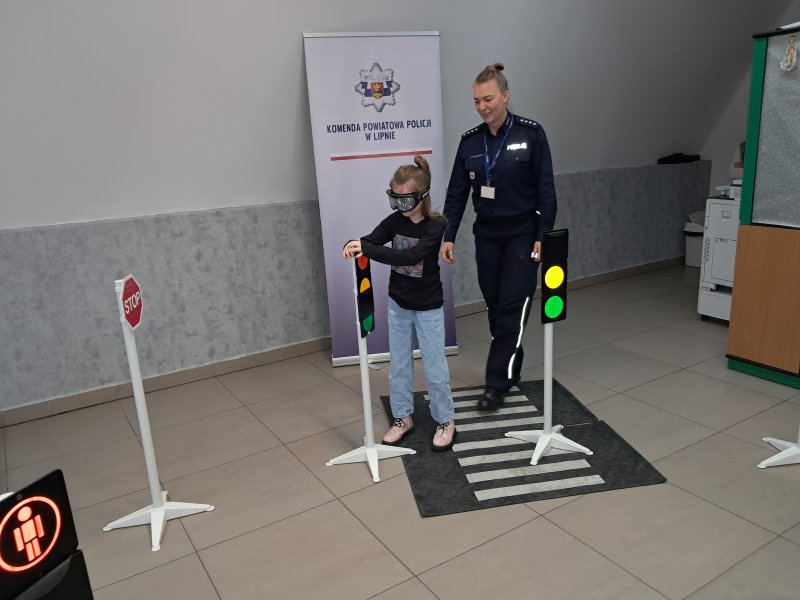 Zdjęcie przedstawia rzecznika prasowego policji Małgorzatę Małkińską i uczestniczkę zajęć w trakcie scenki sytuacyjnej z alkogoglami na zainscenizowanym przejściu dla pieszych w sali, w której odbywały się zajęcia.  