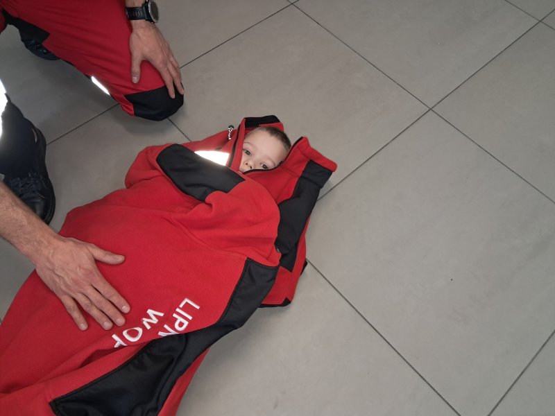 Zdjęcie przedstawia scenkę, w której uczestnik zajęć leży na podłodze i owinięty jest kocem termicznym w celu podniesienia temperatury ciała.  