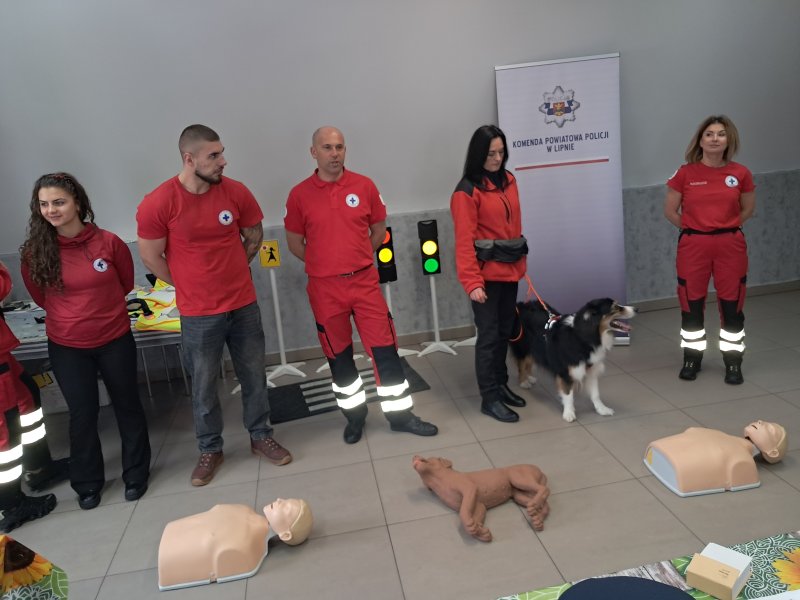 Zdjęcie przedstawia ratowników WOPR z Lipna stojących wraz z psem-ratownikiem w Sali. Przed ratownikami na podłodze leżą fantomy służące do nauki z pierwszej pomocy.  