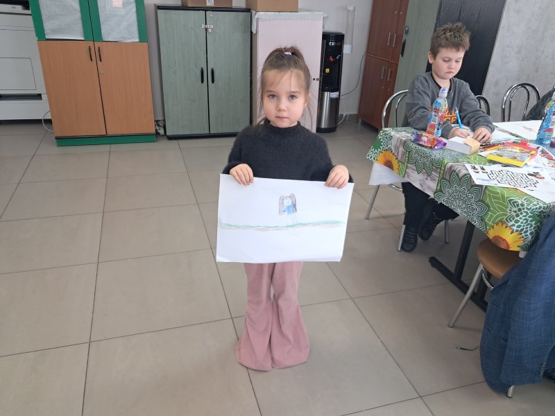 Zdjęcie przedstawia dziewczynkę stojącą i pozującą do zdjęcia z własnym rysunkiem  