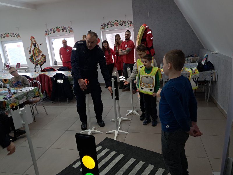 Zdjęcie przedstawia policjantów i uczestnika zajęć w trakcie scenki sytuacyjnej na zainscenizowanym przejściu dla pieszych w sali, w której odbywały się zajęcia. W dali widać pozostałych uczestników zajęć.  