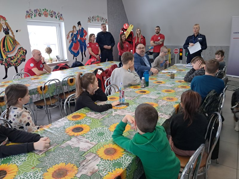 Zdjęcie przedstawia rzecznika prasowego policji Małgorzatę Małkińską stojącą w sali, w której odbywały się zajęcia i mówiącą do dzieci, które siedzą przy stoliku. W dali widać ratowników Lipnowskiego WOPR  