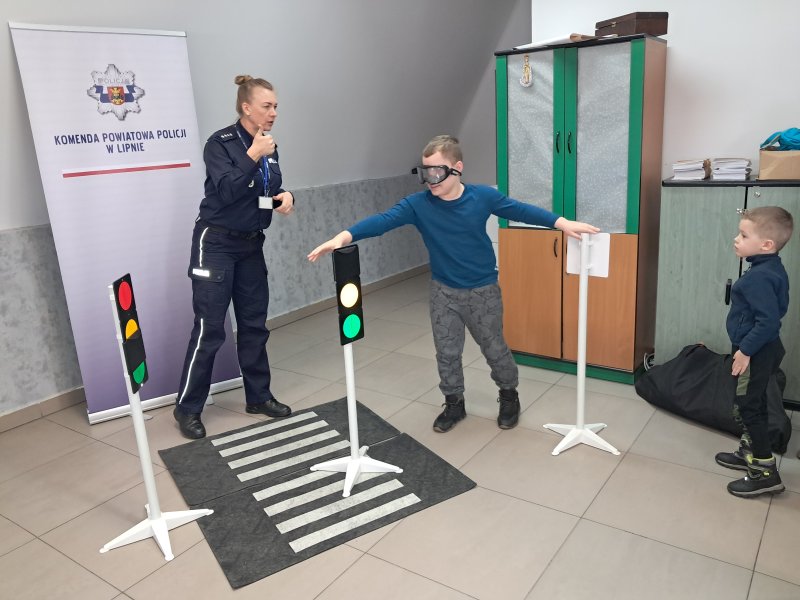 Zdjęcie przedstawia rzecznika prasowego policji Małgorzatę Małkińską i uczestnika zajęć w trakcie scenki sytuacyjnej z alkogoglami na zainscenizowanym przejściu dla pieszych w sali, w której odbywały się zajęcia.  