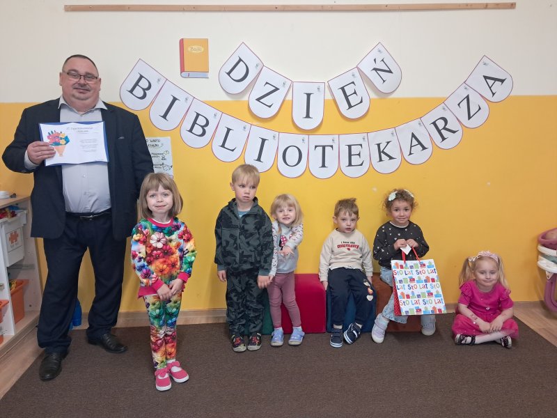 Wspólna fotografia dzieci i dyrektora biblioteki na tle napisu na ścianie „Dzień Bibliotekarza”  