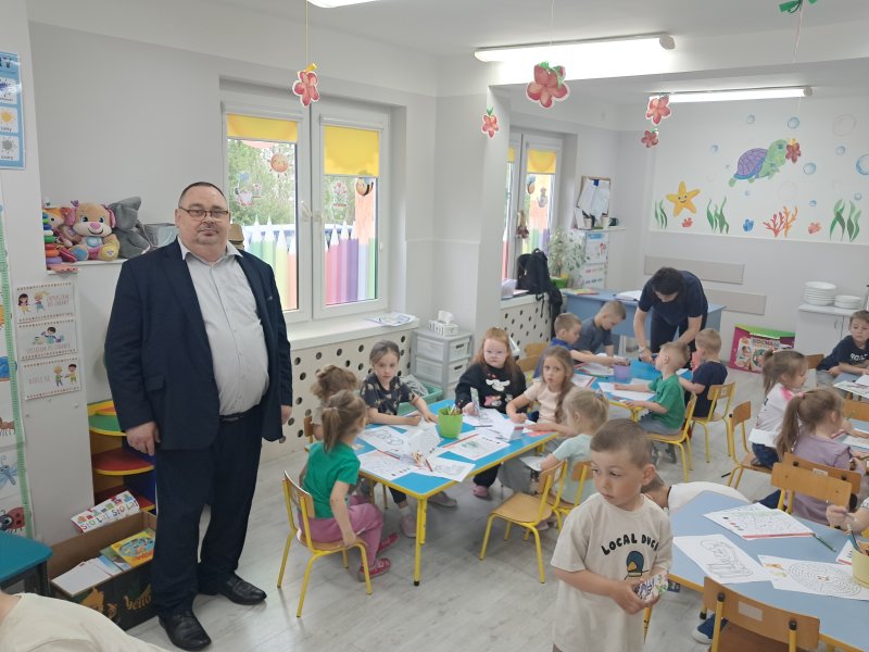 Na zdjęciu widać dyrektora biblioteki w towarzystwie siedzących przy stolikach przedszkolaków w Publicznym Przedszkolu w Skępem  
