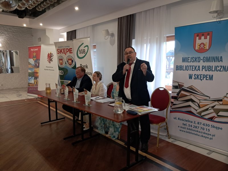 Zdjęcie przedstawia jurorów siedzących przy stoliku. Przewodniczący Jury stoi i przemawia przez mikrofon  