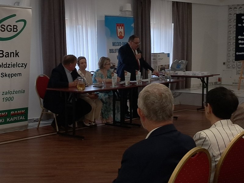 Zdjęcie przedstawia jurorów siedzących przy stoliku. Przewodniczący Jury stoi i przemawia przez mikrofon  