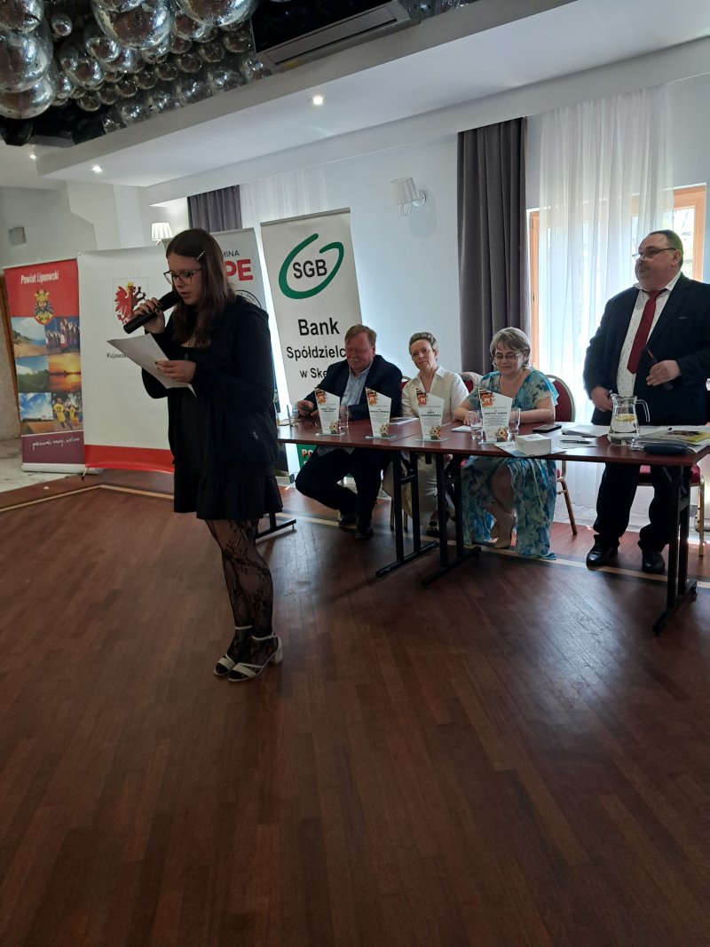 Zdjęcie przedstawia laureatkę konkursu czytającą nagrodzony wiersz przy stole Jurorów konkursu  