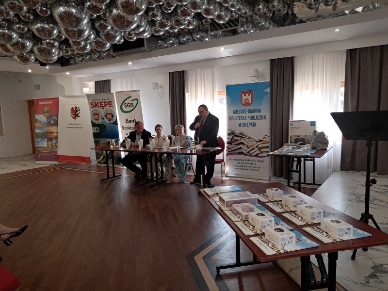 Zdjęcie przedstawia jurorów siedzących przy stoliku. Przewodniczący Jury stoi i przemawia przez mikrofon  